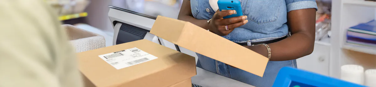 ParcelShop store assistant is smiling and using a blue hand-held device to scan multiple parcels placed on the countertop in front of them. The customer can be seen in the foreground.