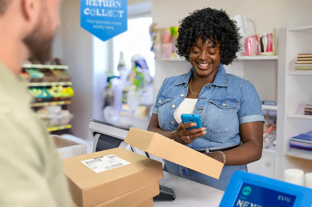 ParcelShop store assistant is smiling and using a blue hand-held device to scan multiple parcels placed on the countertop in front of them. The customer can be seen in the foreground.