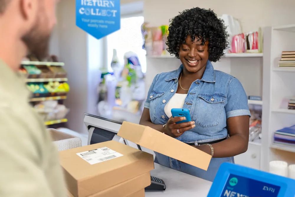 ParcelShop store assistant is smiling and using a blue hand-held device to scan multiple parcels placed on the countertop in front of them. The customer can be seen in the foreground.