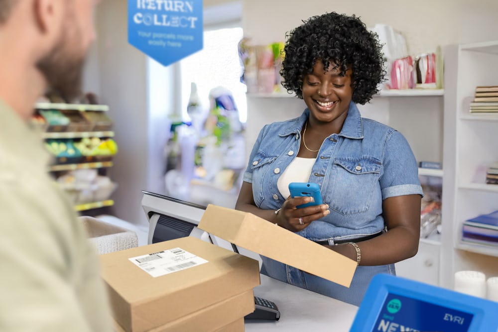 ParcelShop store assistant is smiling and using a blue hand-held device to scan multiple parcels placed on the countertop in front of them. The customer can be seen in the foreground.