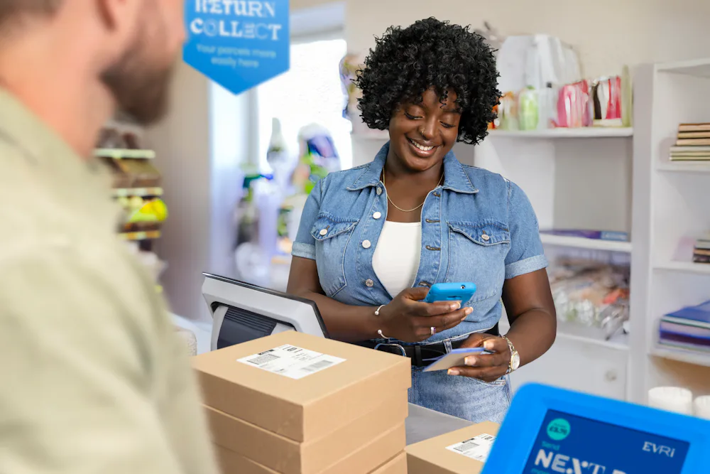 ParcelShop store assistant is smiling and holding a blue hand-held device to scan a ParcelShop receipt to confirm the parcels have been dropped off.