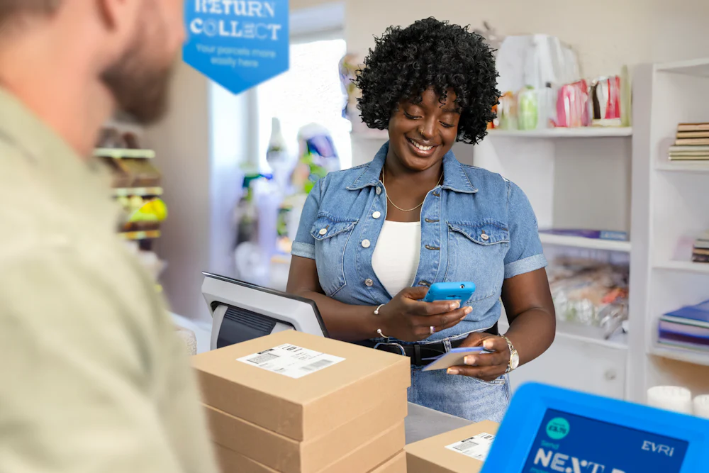 ParcelShop store assistant is smiling and holding a blue hand-held device to scan a ParcelShop receipt to confirm the parcels have been dropped off.