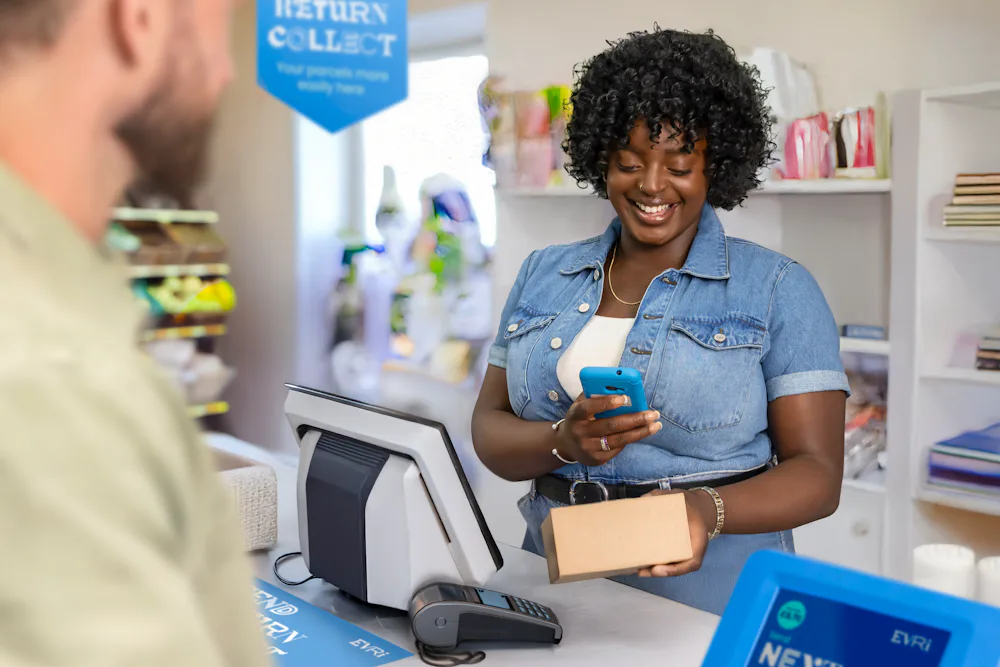 ParcelShop store assistant is smiling and using a blue hand-held device to scan a parcel. The customer can be seen in the foreground.
