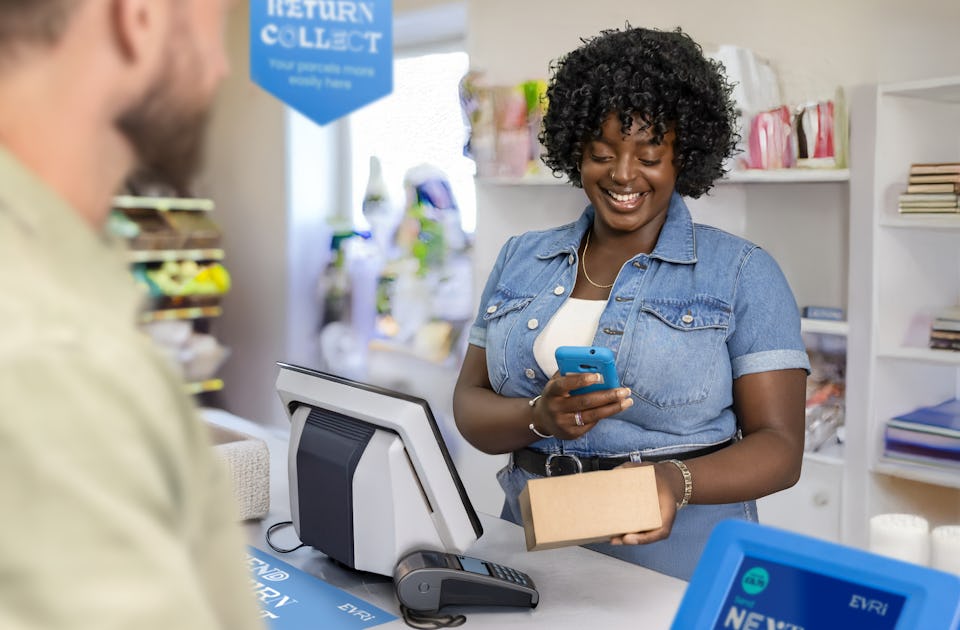 ParcelShop store assistant is smiling and using a blue hand-held device to scan a parcel. The customer can be seen in the foreground.
