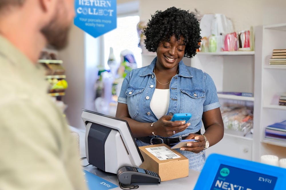 ParcelShop store assistant is smiling and holding a blue hand-held device to scan a ParcelShop receipt to confirm the parcels have been dropped off. The customer can be seen in the foreground.