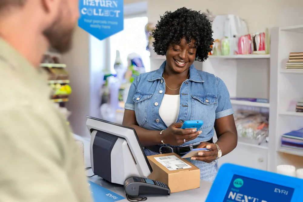 ParcelShop store assistant is smiling and holding a blue hand-held device to scan a ParcelShop receipt to confirm the parcels have been dropped off. The customer can be seen in the foreground.