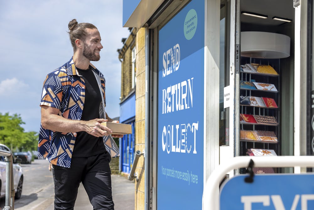 Customer stood outside of a ParcelShop store smiling and holding a parcel. The blue Evri signage can be seen on the storefront.