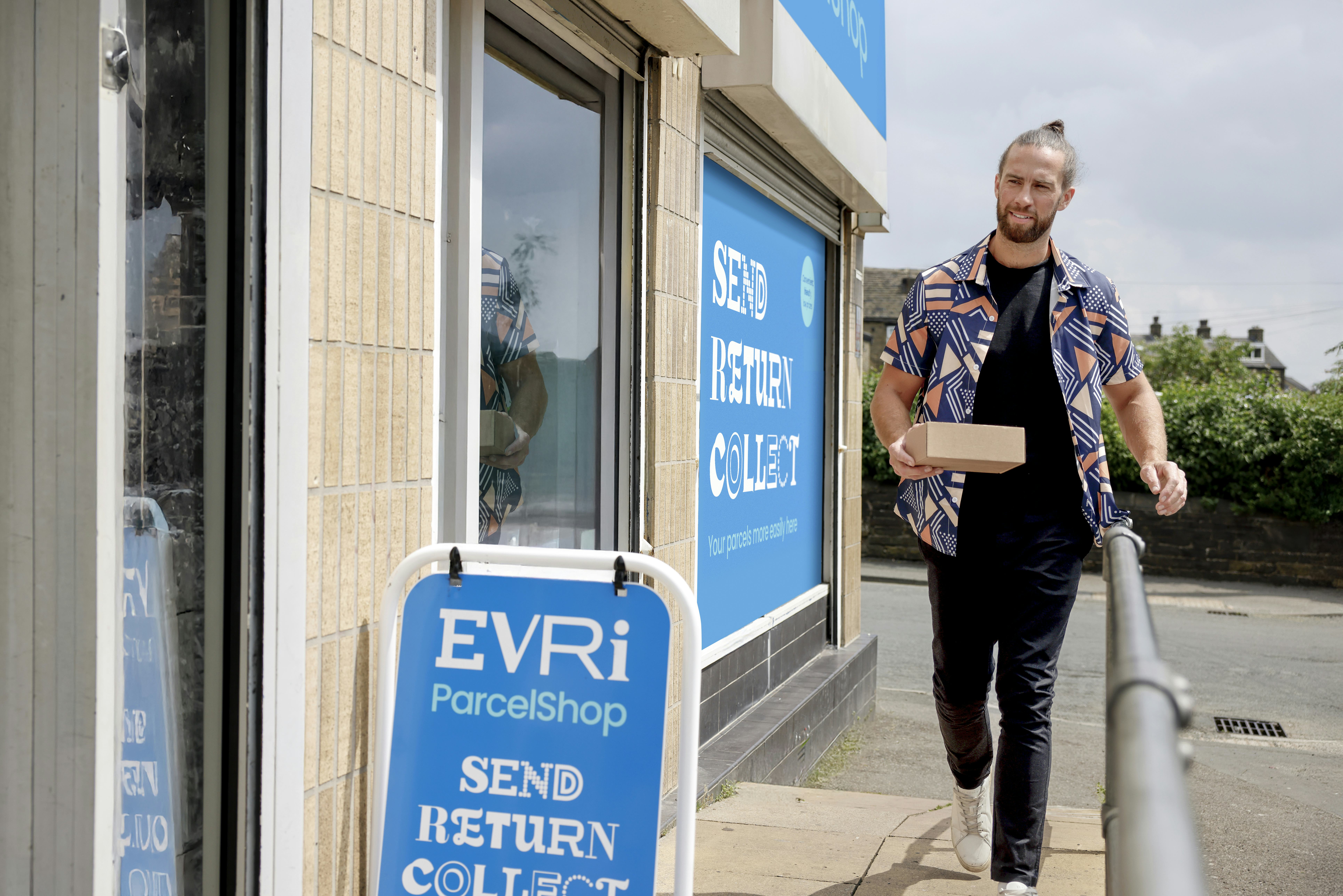 Customer walking towards a ParcelShop store holding a parcel. The blue Evri signage can be seen on the storefront with a free-standing sign near the entrance.