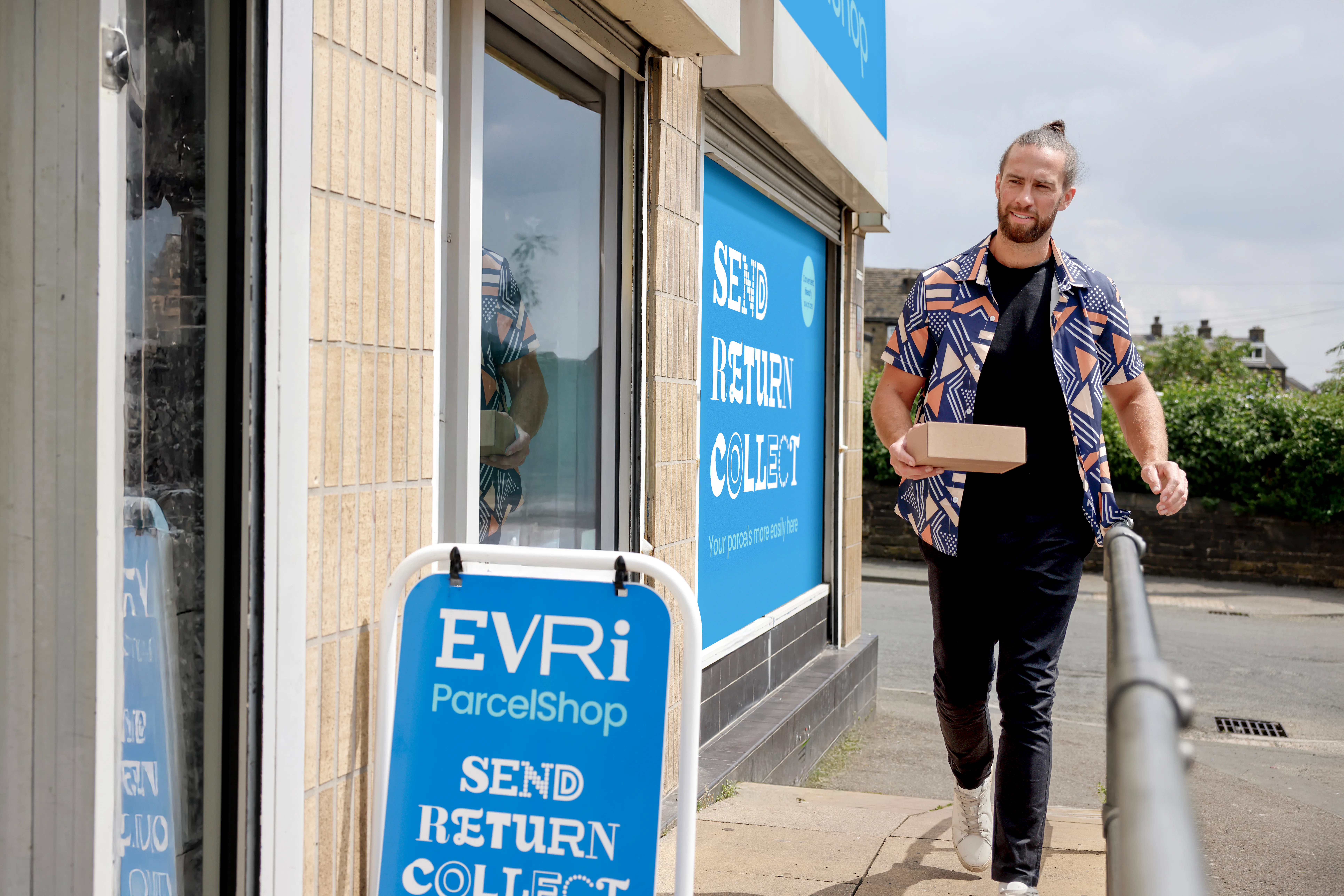 Customer walking towards a ParcelShop store holding a parcel. The blue Evri signage can be seen on the storefront with a free-standing sign near the entrance.