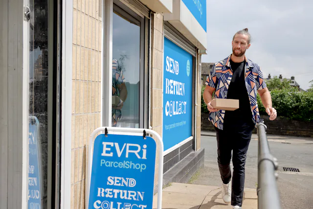 Customer walking towards a ParcelShop store holding a parcel. The blue Evri signage can be seen on the storefront with a free-standing sign near the entrance.
