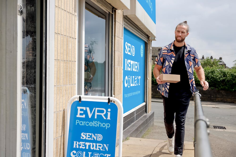 Customer walking towards a ParcelShop store holding a parcel. The blue Evri signage can be seen on the storefront with a free-standing sign near the entrance.