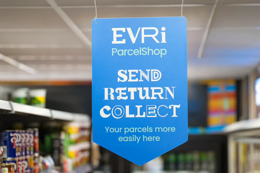 Evri blue ParcelShop sign hung from the ceiling of a store in one of the aisles. Stock on shelves is visible in the background.