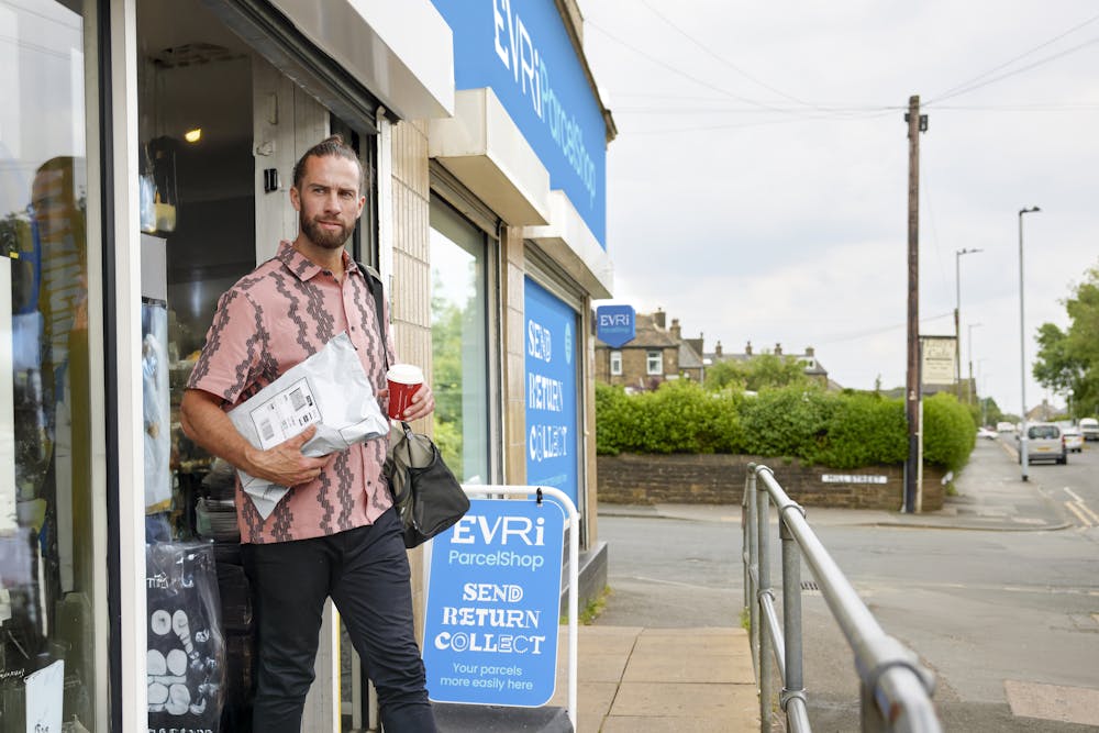 Customer is walking away from a ParcelShop, holding their collected parcel in one hand and a store-bought cup of coffee in the other hand.