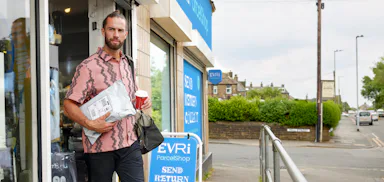Customer is walking away from a ParcelShop, holding their collected parcel in one hand and a store-bought cup of coffee in the other hand.
