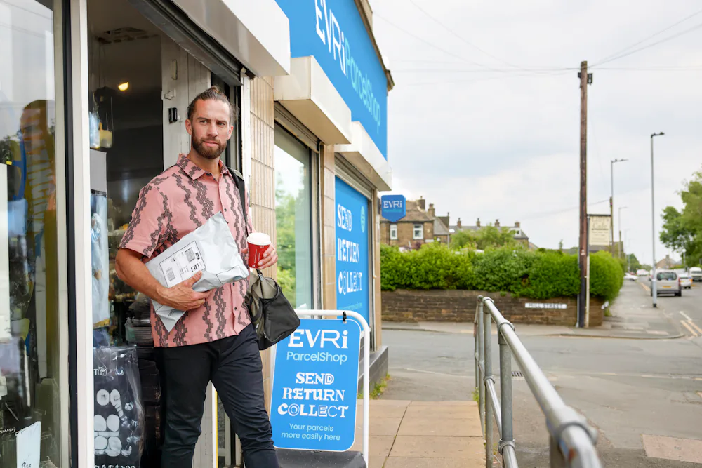 Customer is walking away from a ParcelShop, holding their collected parcel in one hand and a store-bought cup of coffee in the other hand.