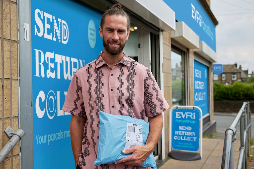 Customer stood outside of a ParcelShop store smiling and holding a parcel. The blue Evri signage can be seen on the storefront.