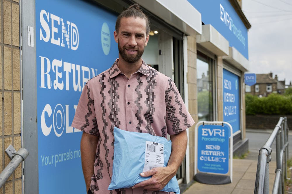 Customer stood outside of a ParcelShop store smiling and holding a parcel. The blue Evri signage can be seen on the storefront.