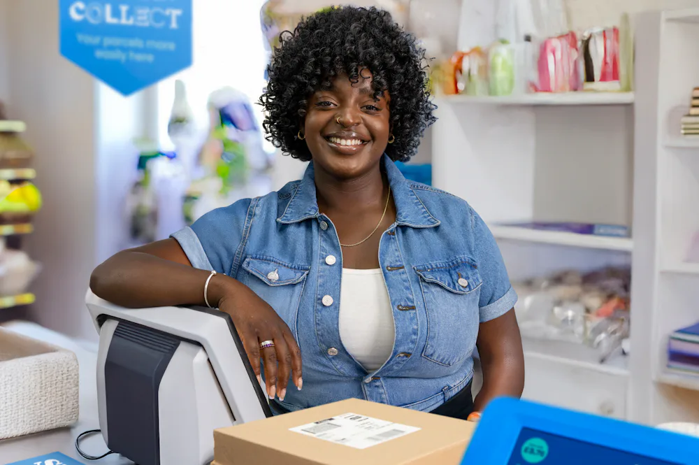 ParcelShop store assistant is standing at the till and smiling, posing for the photo.