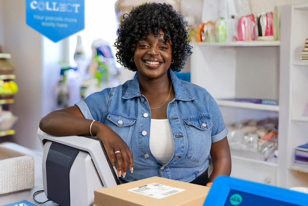ParcelShop store assistant is standing at the till and smiling, posing for the photo.