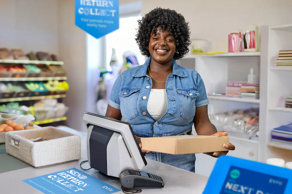 ParcelShop store assistant is standing at the till, holding a parcel and smiling for the photo.