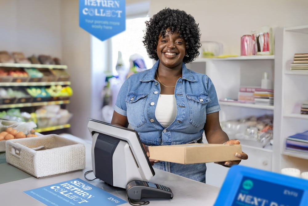 ParcelShop store assistant is standing at the till, holding a parcel and smiling for the photo.