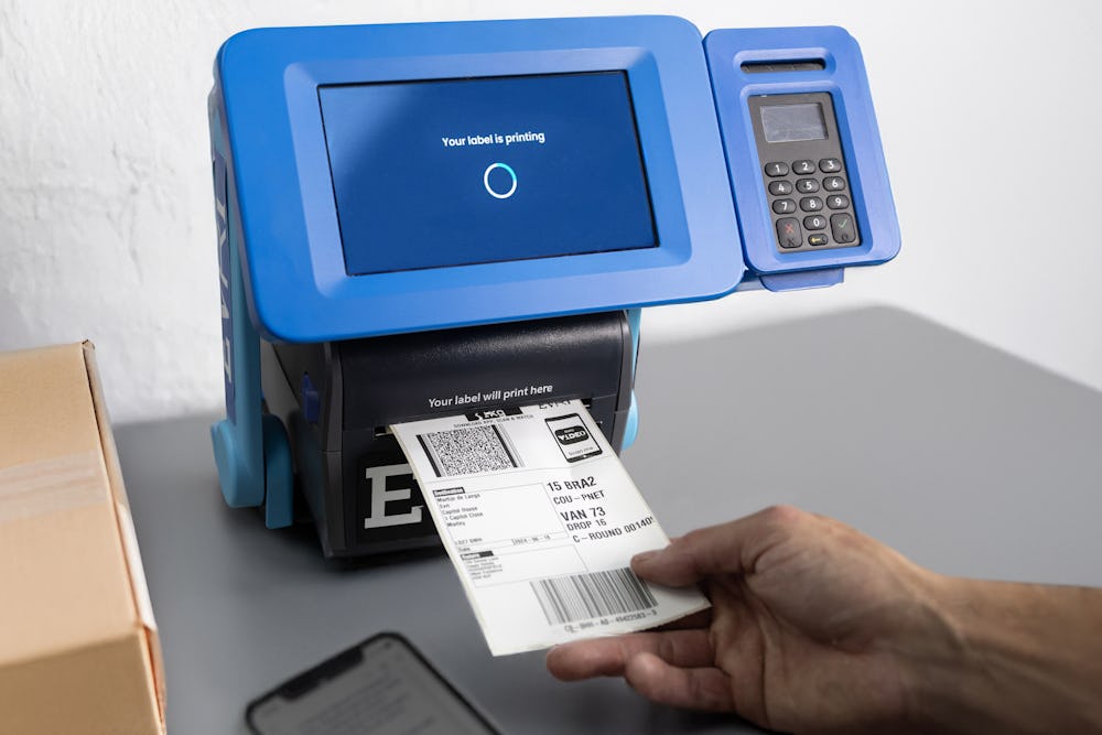 A close-up of a ParcelShop Print in Store device printing a label. A hand is holding the label, ready to tear off and apply to the parcel.