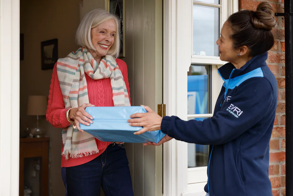 A female courier hands a parcel to a recipient. The recipient, an older woman, is smiling.