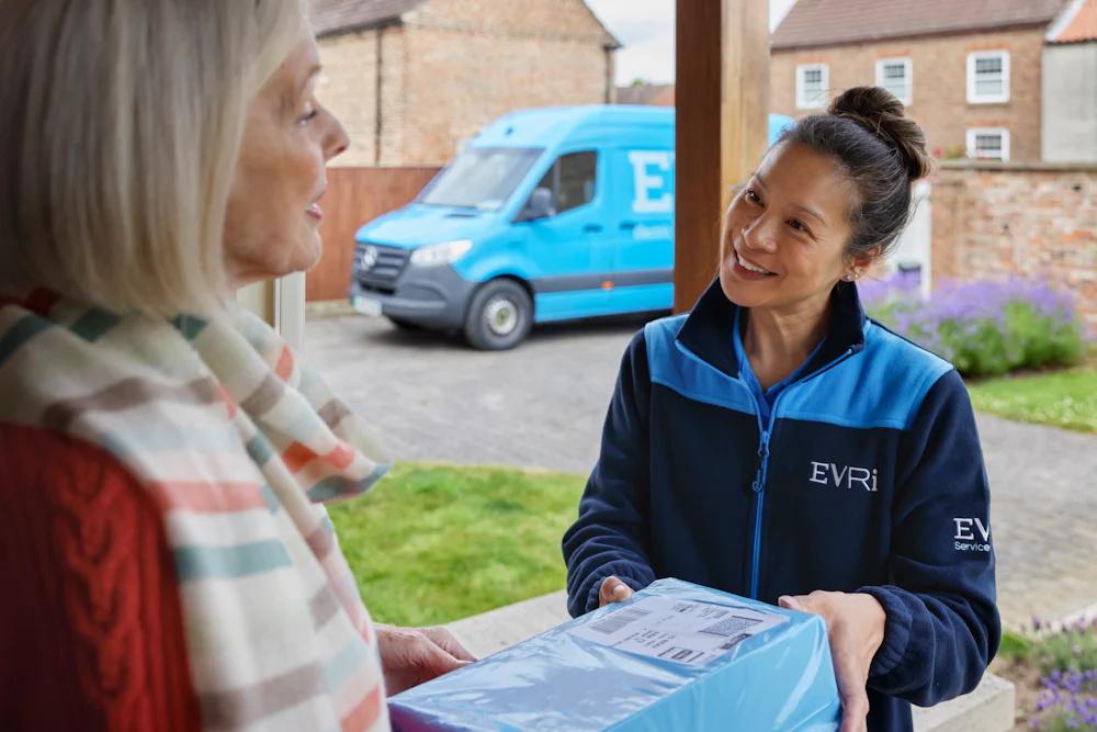 A female courier hands a parcel to a recipient. A blue Evri van can be seen in the background.