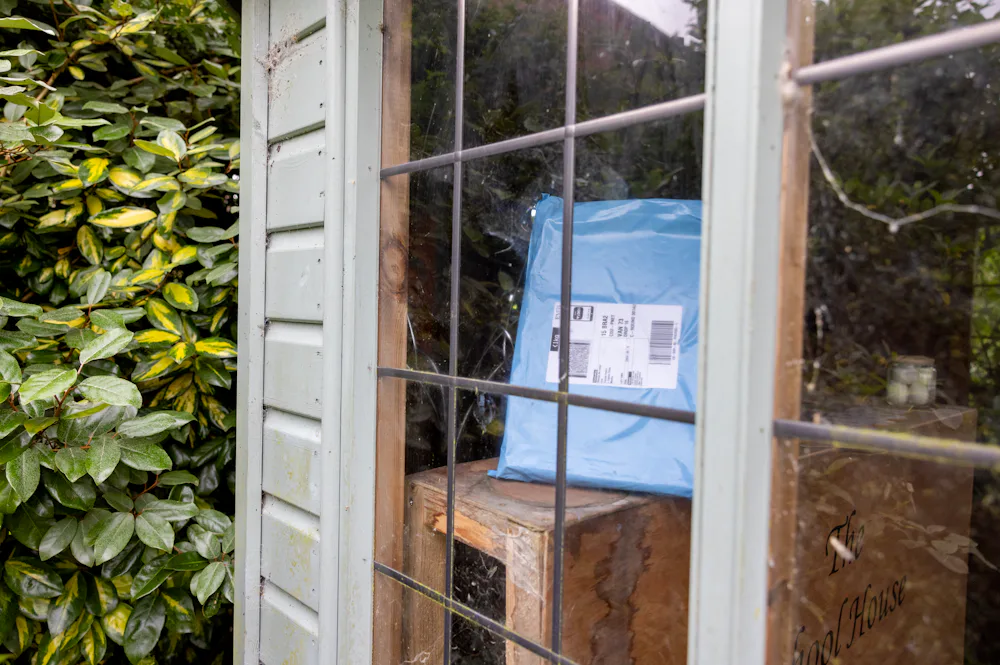 A parcel can be seen through the window of a summer house, safely placed inside out of public view and protected from the elements. A hedge is in the background, behind the summer house.