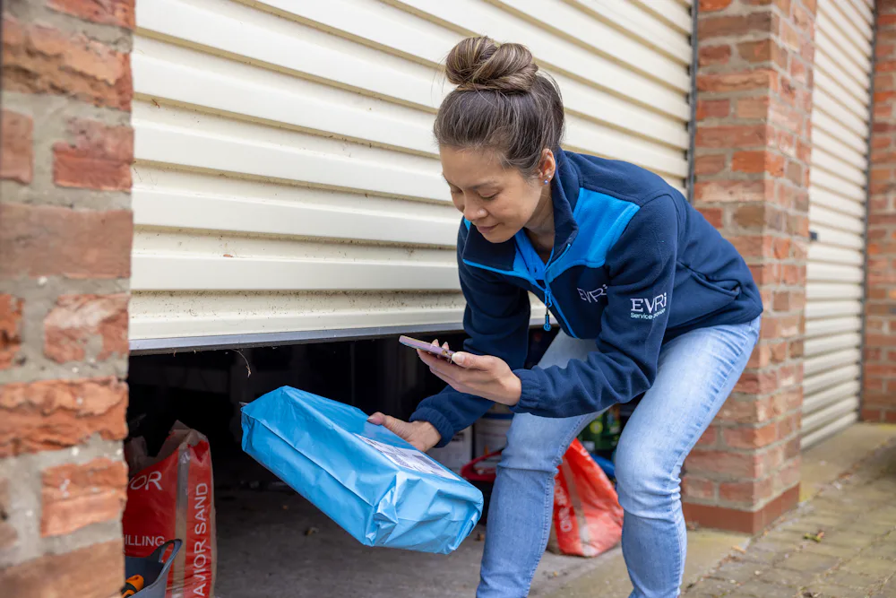 A courier is outside next to an open garage door, scanning a parcel for its delivery to a safe place.