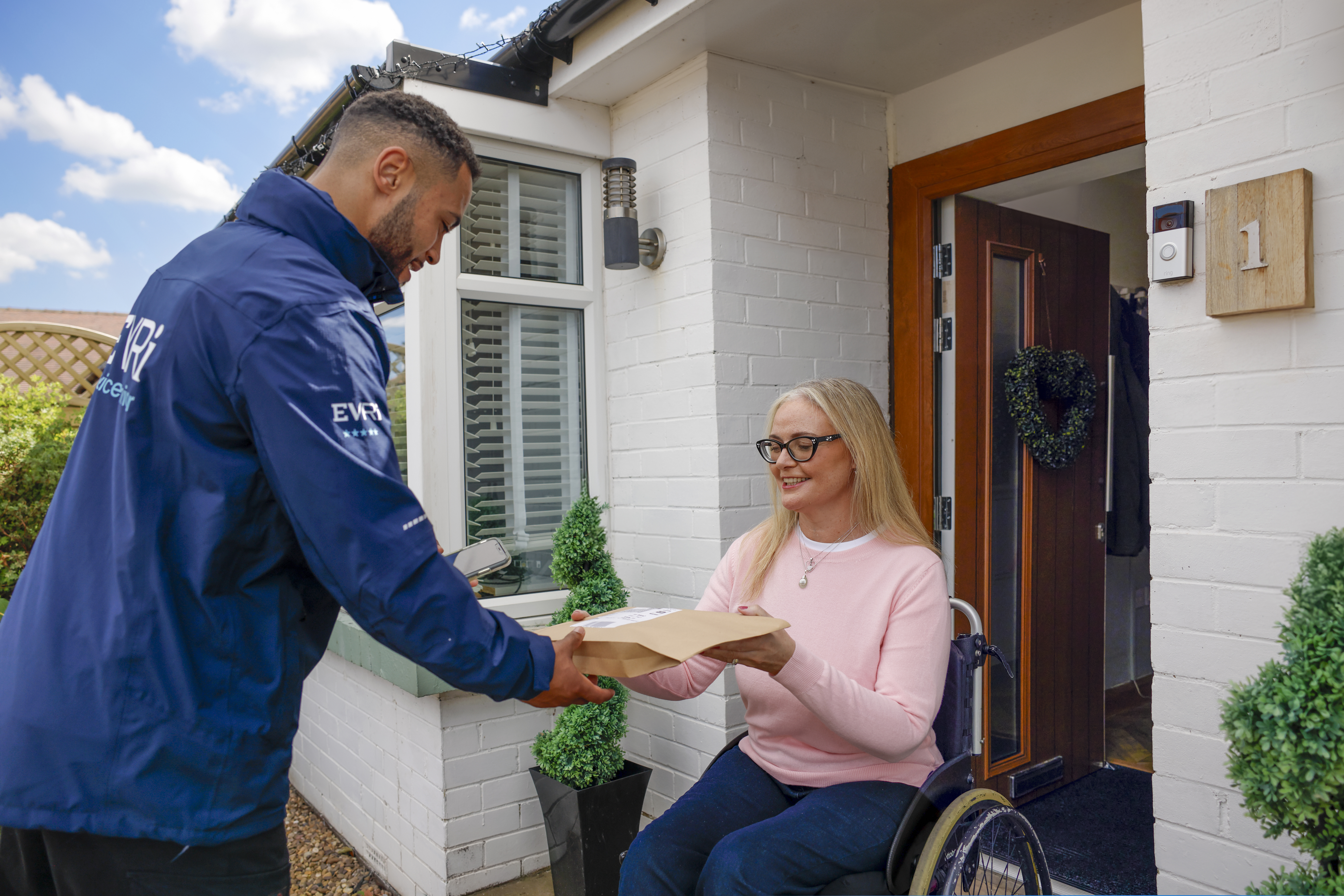 A customer sitting in a wheelchair is outside their house and accepting their parcel from the courier. The customer is in full view with a side profile of the courier.