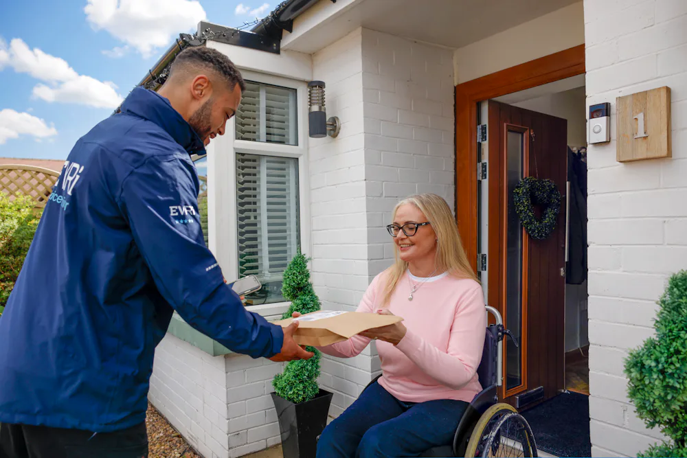 A customer sitting in a wheelchair is outside their house and accepting their parcel from the courier. The customer is in full view with a side profile of the courier.