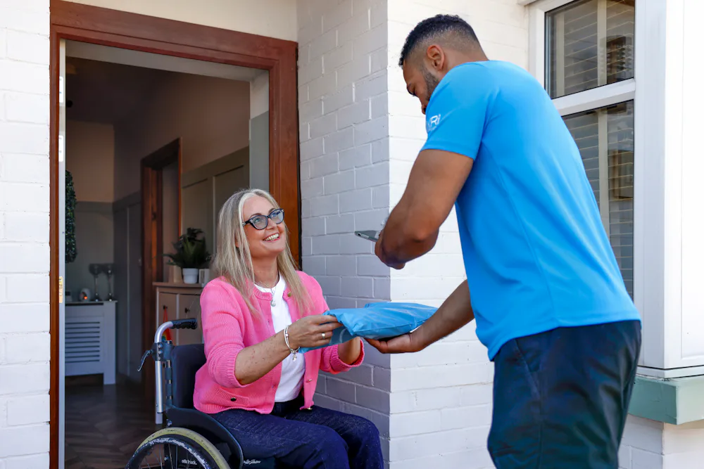 A customer sat in a wheelchair is outside their front door smiling as the courier hands a parcel to them. The courier is taking a photo of the parcel being received as proof of delivery.