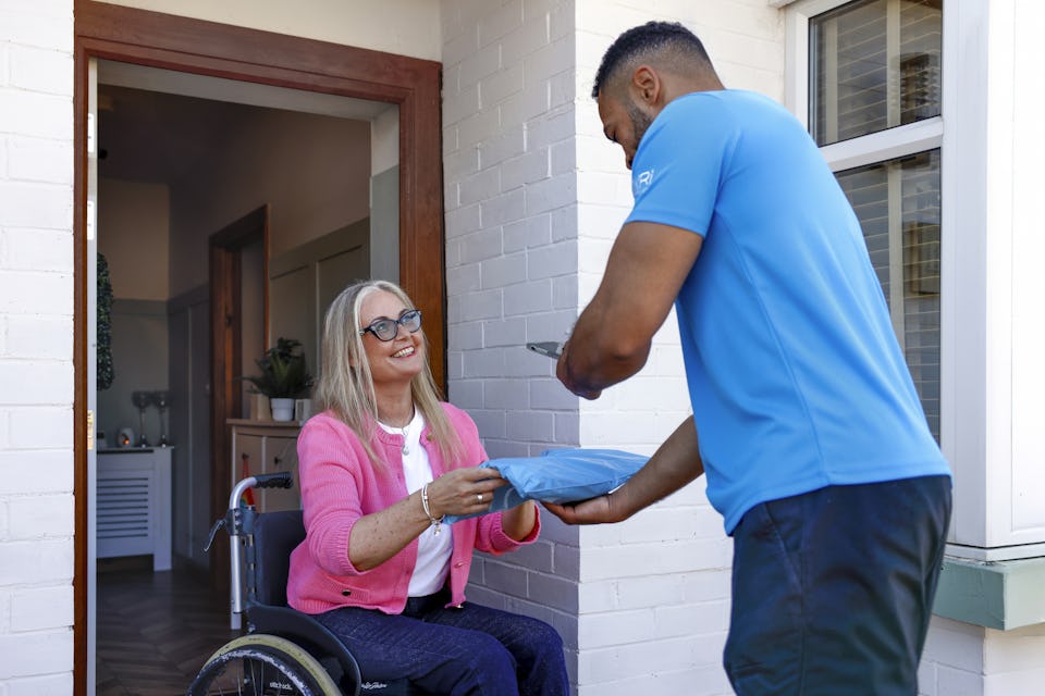 A customer sat in a wheelchair is outside their front door smiling as the courier hands a parcel to them. The courier is taking a photo of the parcel being received as proof of delivery.