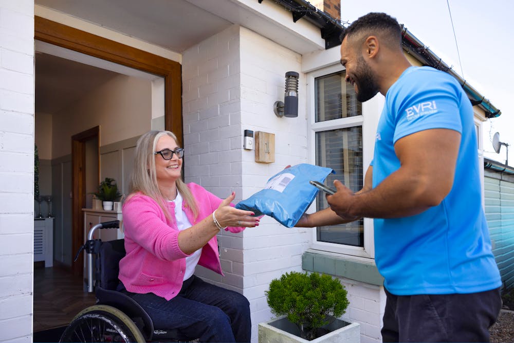 A customer sitting in a wheelchair is outside their front door smiling as the courier hands a parcel to them.