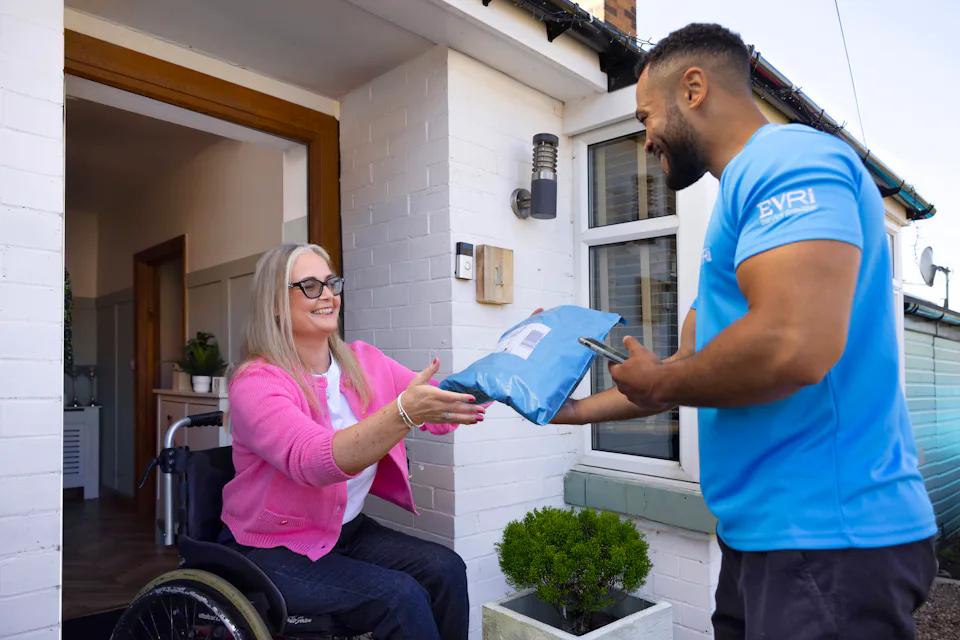 A customer sitting in a wheelchair is outside their front door smiling as the courier hands a parcel to them.