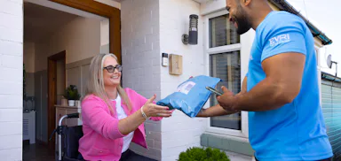 A customer sitting in a wheelchair is outside their front door smiling as the courier hands a parcel to them.