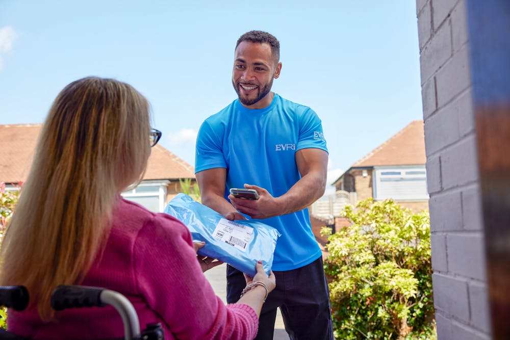 A customer sitting in a wheelchair is outside and a courier is handing a parcel to them with a smile. The customer is positioned to be facing away from the camera, with the courier in full view.
