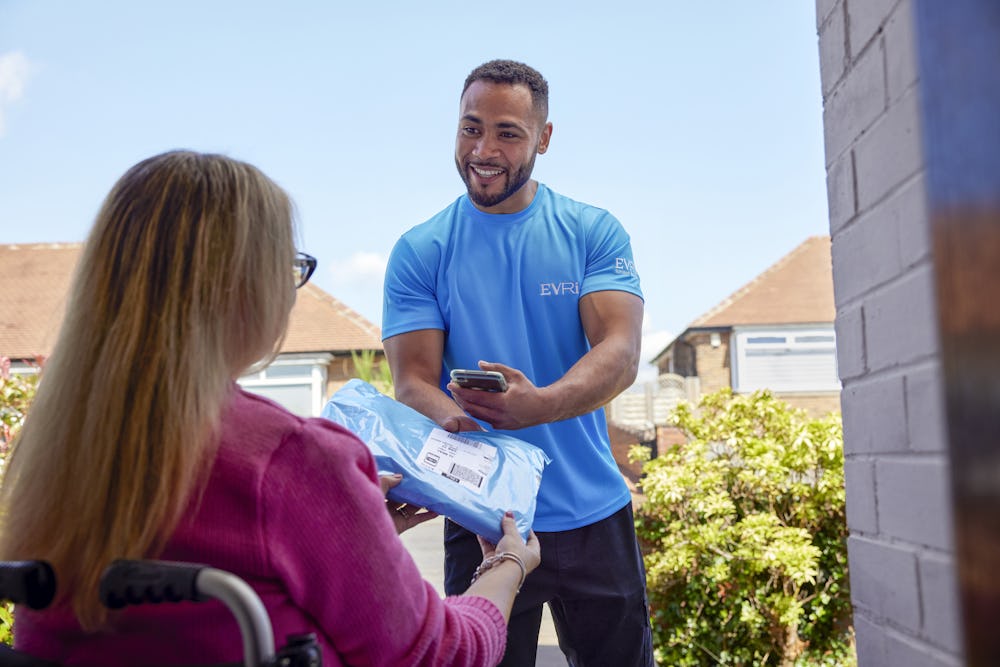 A customer sitting in a wheelchair is outside and a courier is handing a parcel to them with a smile. The customer is positioned to be facing away from the camera, with the courier in full view.
