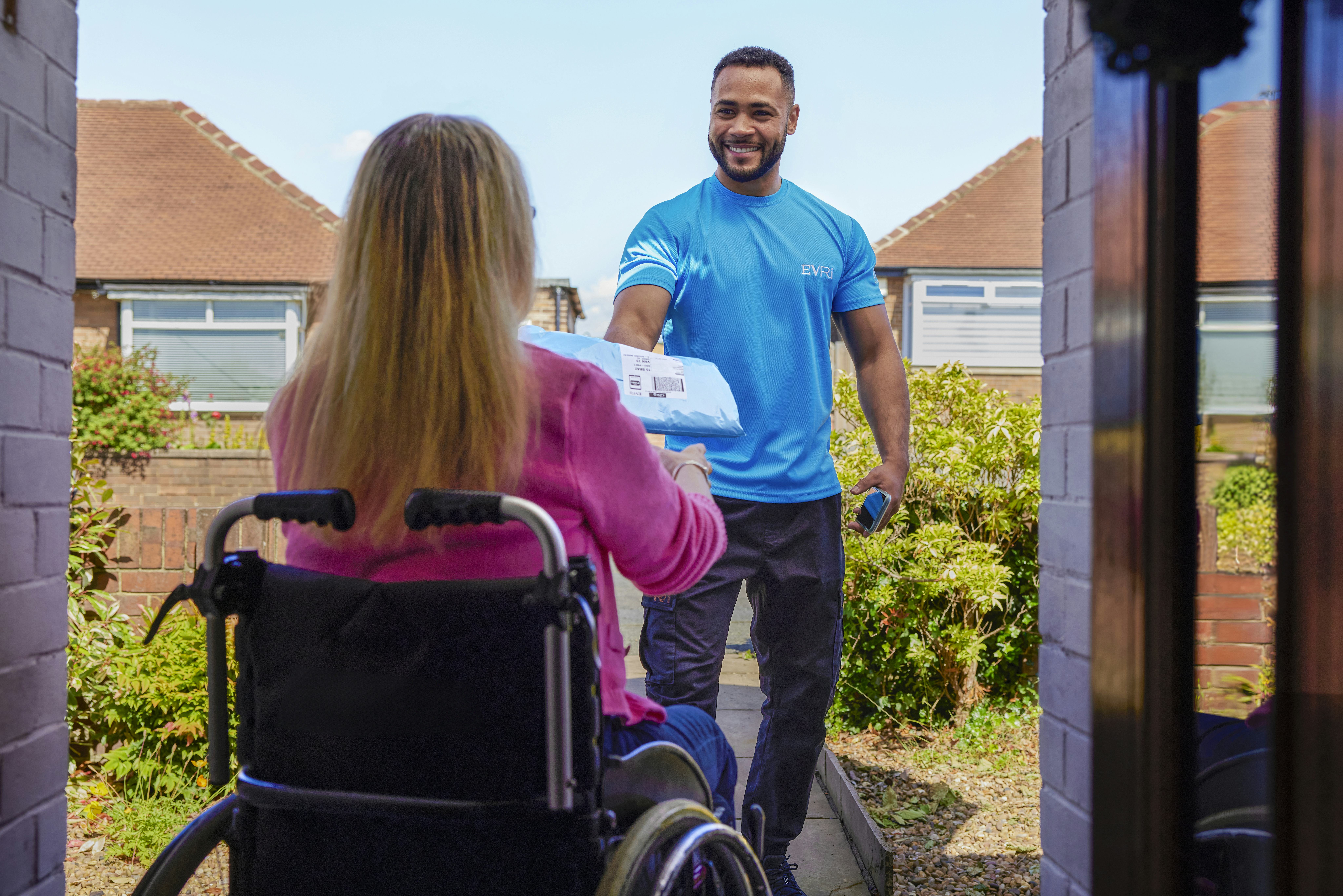 A customer sitting in a wheelchair is outside and a courier is handing a parcel to them with a smile. The customer is positioned to be facing away from the camera, with the courier in full view.