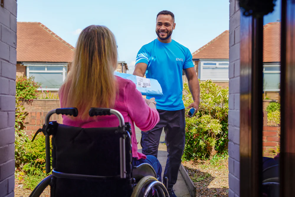 A customer sitting in a wheelchair is outside and a courier is handing a parcel to them with a smile. The customer is positioned to be facing away from the camera, with the courier in full view.