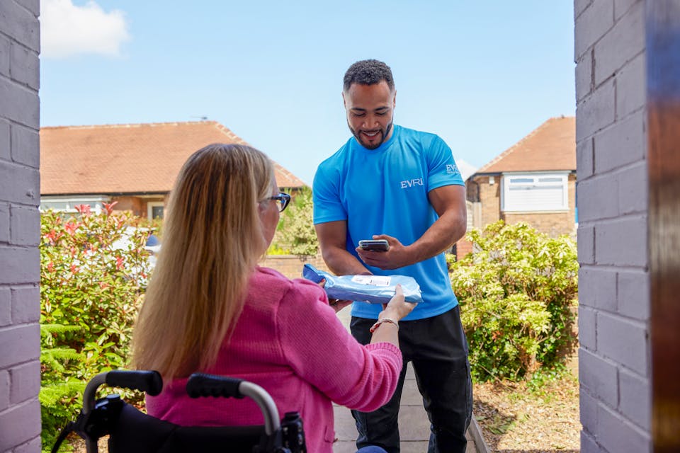 A customer sitting in a wheelchair is outside and a courier is handing a parcel to them with one hand and using a device in the other hand to scan the parcel. The customer is positioned to be facing away from the camera, with the courier in full view.