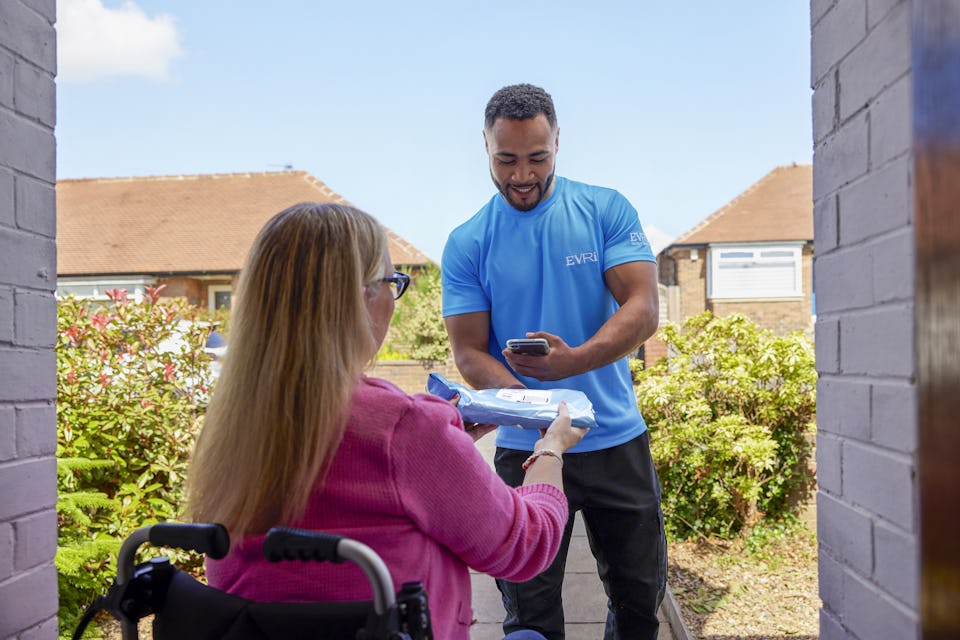 A customer sitting in a wheelchair is outside and a courier is handing a parcel to them with one hand and using a device in the other hand to scan the parcel. The customer is positioned to be facing away from the camera, with the courier in full view.