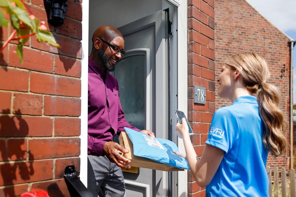 A customer is standing in the doorway of their home smiling and holding onto parcels. A courier is standing opposite on the top step smiling and taking a photo of the parcels for proof of delivery.