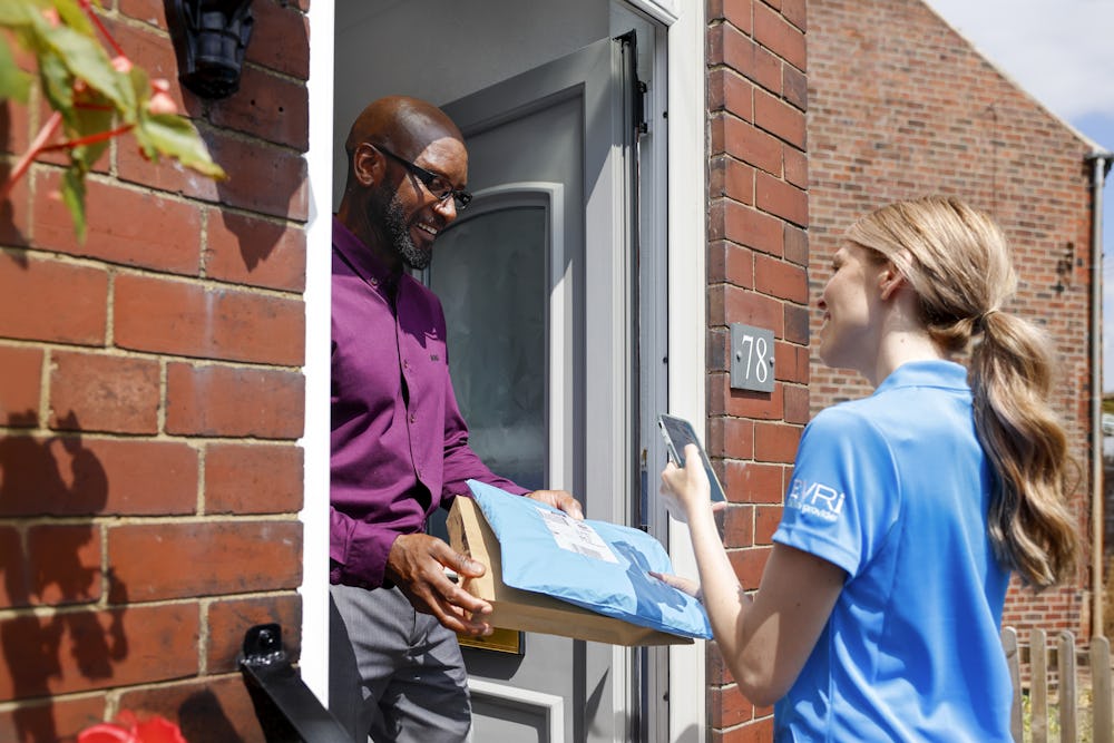 A customer is standing in the doorway of their home smiling and holding onto parcels. A courier is standing opposite on the top step smiling and taking a photo of the parcels for proof of delivery.
