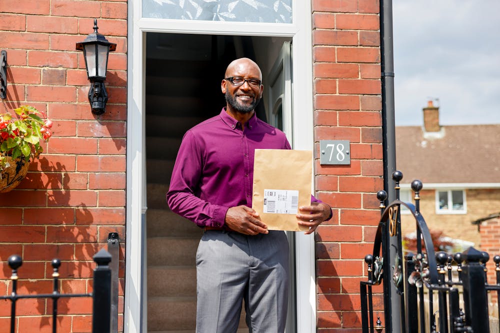 A customer is standing in the doorway of their home holding their parcel and smiling for the photo.