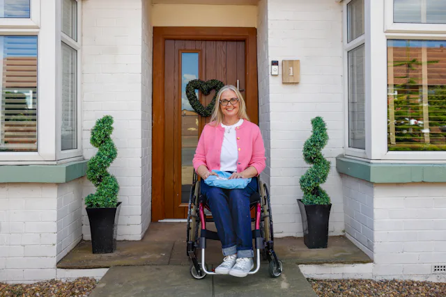 A customer is outside their home and sitting in their wheelchair, smiling for the photo with a parcel on their lap.