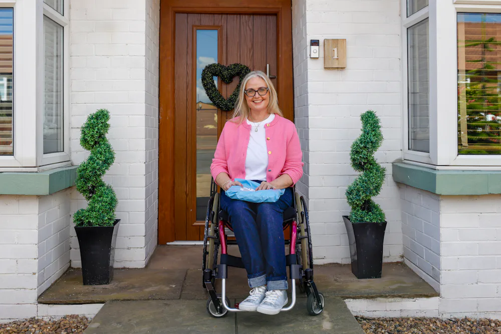 A customer is outside their home and sitting in their wheelchair, smiling for the photo with a parcel on their lap.