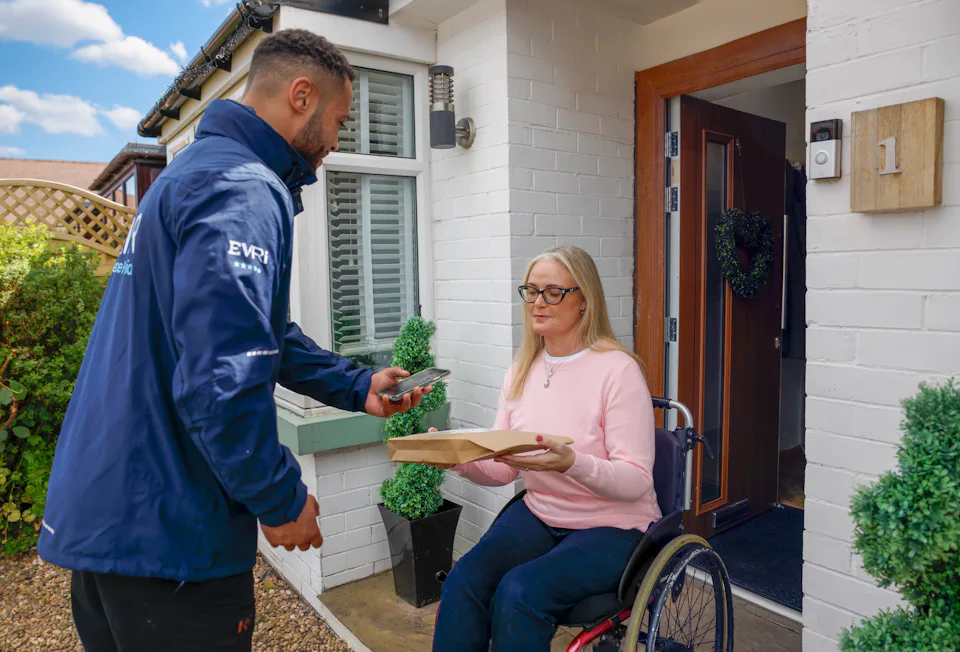 A customer sitting in a wheelchair is outside their house and holding their parcel as the courier takes a photo for proof of delivery. The customer is in full view with a side profile of the courier.