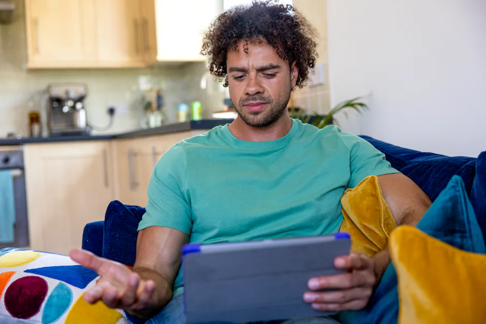 A customer sat on a sofa, holding their tablet and looking frustratingly at their screen.